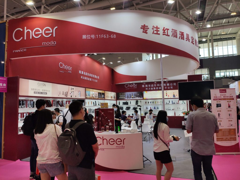 Attendees browse a red and white exhibition booth displaying numerous products on shelves. A large curved sign hangs above the busy trade show floor.