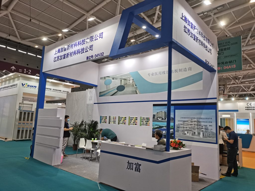 Exhibition booth in a busy convention hall. A woman stands behind a desk while a man examines product samples. Other attendees are in the background.
