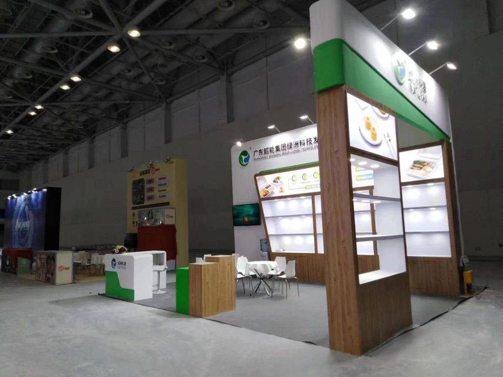 Wide shot of an empty trade show booth featuring white, green, and wood-look panels, shelves, and a reception desk in a large hall.