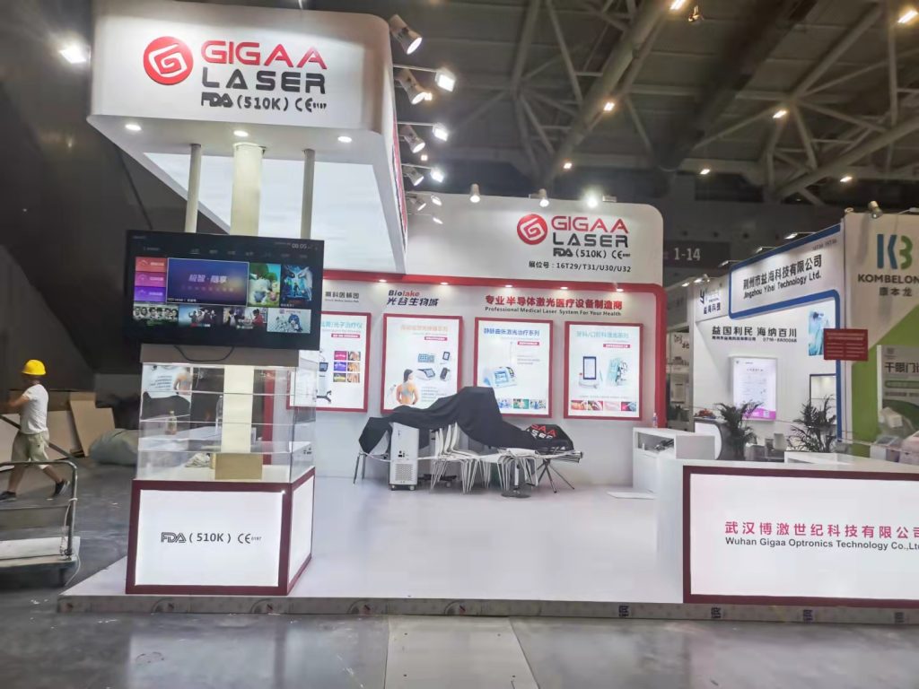 An exhibition booth in a large hall, featuring a large screen, covered display tables, and white walls with red trim, still under construction.
