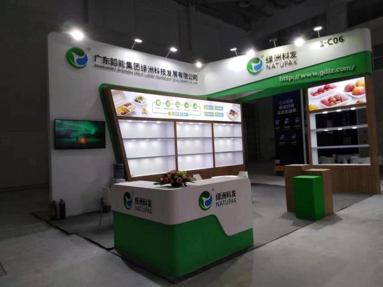 A completed white and green trade show booth with a reception desk, floral arrangement, food displays, and a screen showing an abstract image.