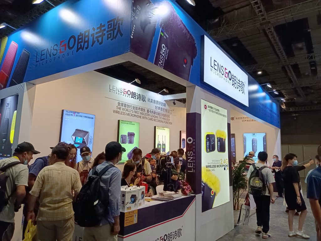 A crowded exhibition booth with many masked attendees looking at microphone displays on illuminated panels and tables.