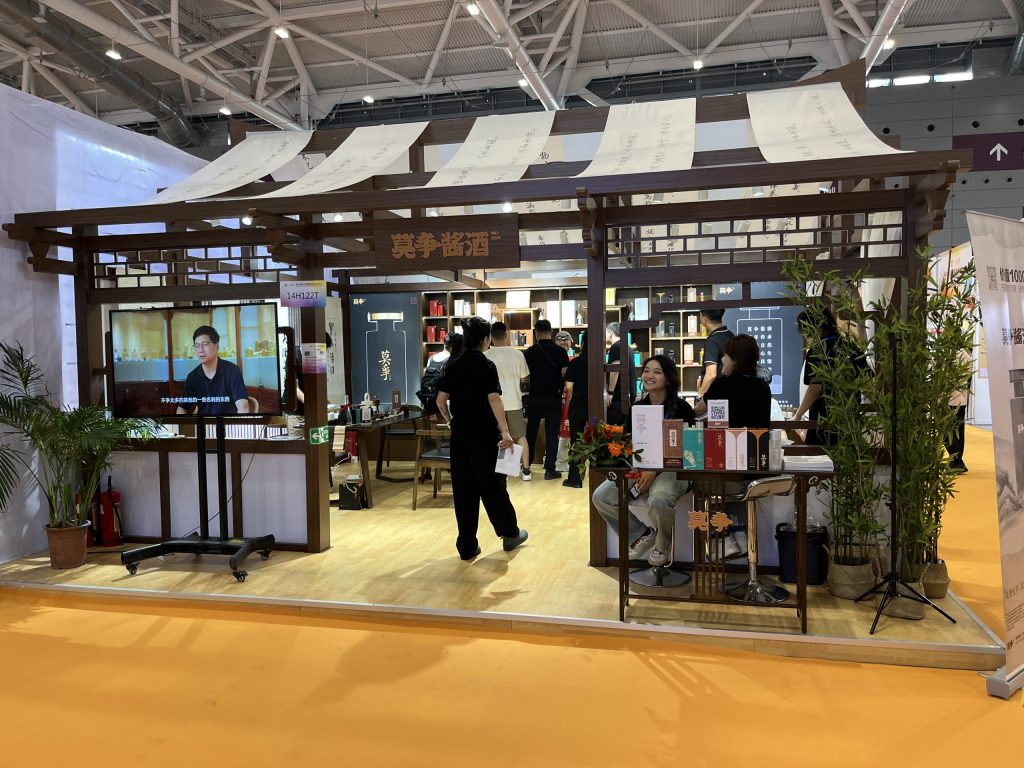 Exhibition booth with traditional East Asian wooden design, people interacting inside, and a large video screen displaying a person.