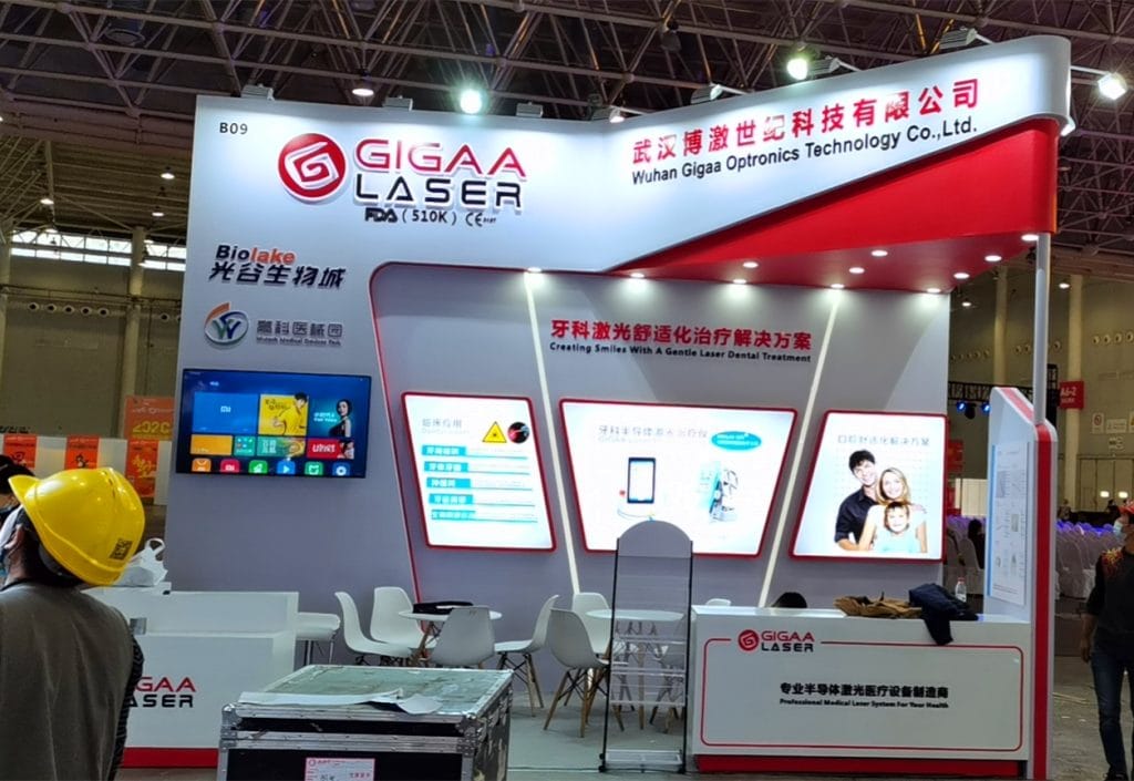 Two photos of an active red and white exhibition booth with digital screens, visitors, and staff members present.