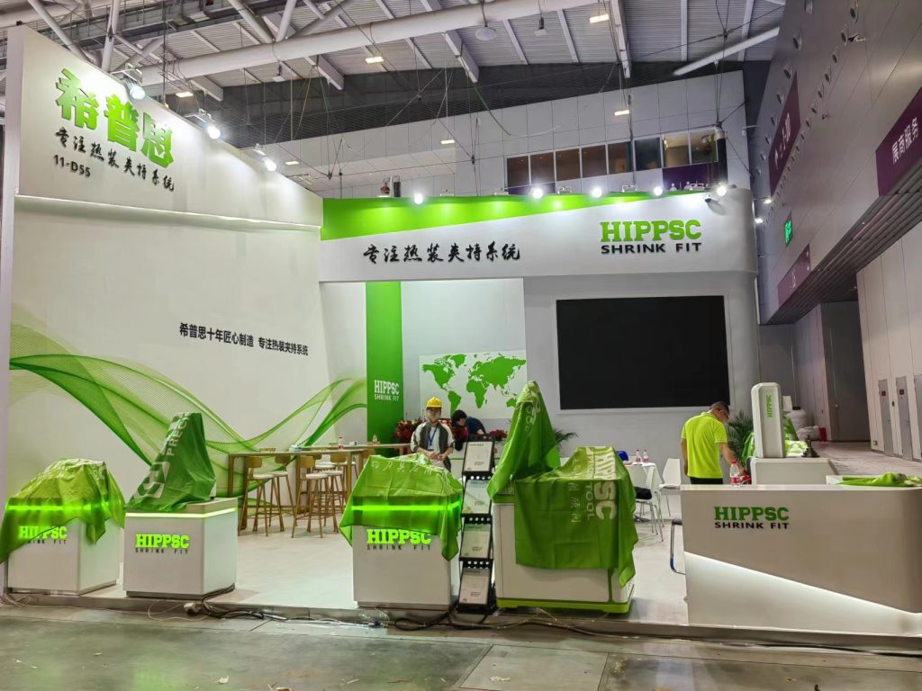 Exhibition booth under construction, with display pedestals covered in green fabric. A worker in a hard hat stands near a table, and another worker is visible in a green shirt.