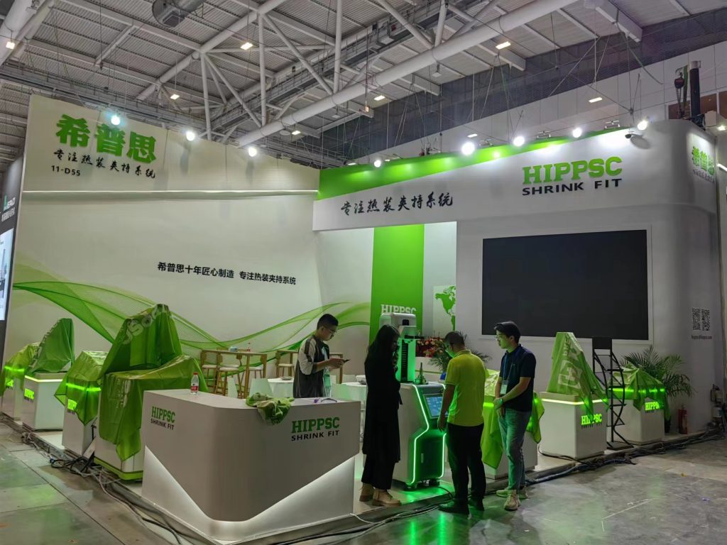 People interact at a white and green exhibition booth with multiple displays. A group of individuals gather near a counter, with products visible on pedestals.
