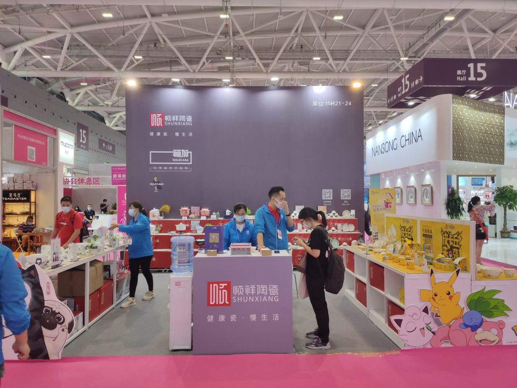 Busy trade show booth. People browse kitchenware and dishes. Booth has a dark back wall and a yellow display with characters.