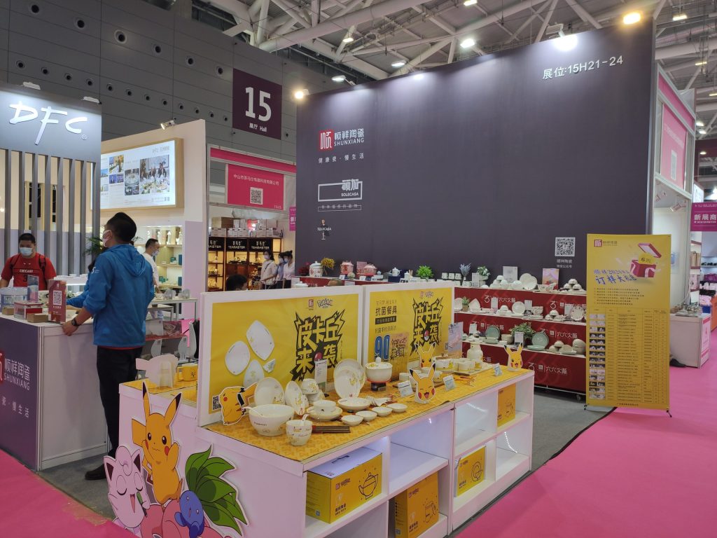 Close-up of a trade show booth's yellow display, featuring character-themed dishes and teapots. Two men stand at a counter.