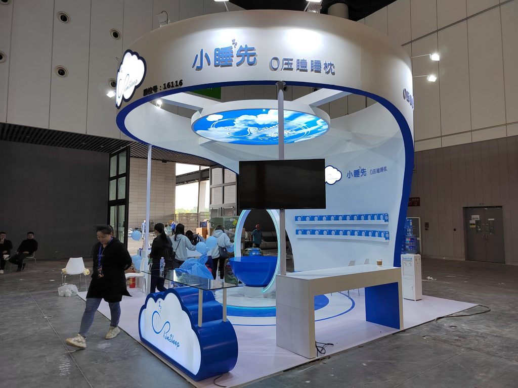 People working at a partially set-up white and blue trade show booth with cloud designs, a TV screen, and displayed products.