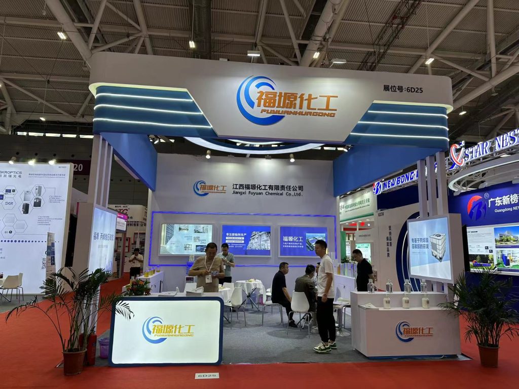 People gather in a white and blue modern exhibition booth at a trade show, with multiple display screens, a reception counter, and a small seating area.
