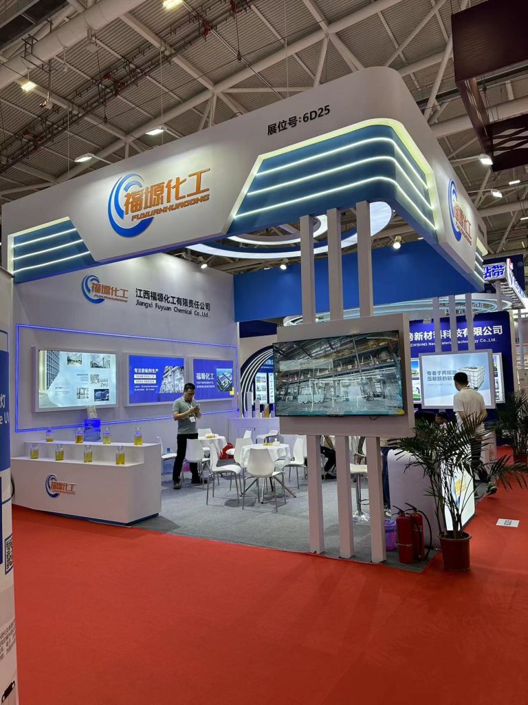 An active exhibition booth with white and blue design elements, featuring display screens, a reception area, and people interacting. Red carpet in foreground.