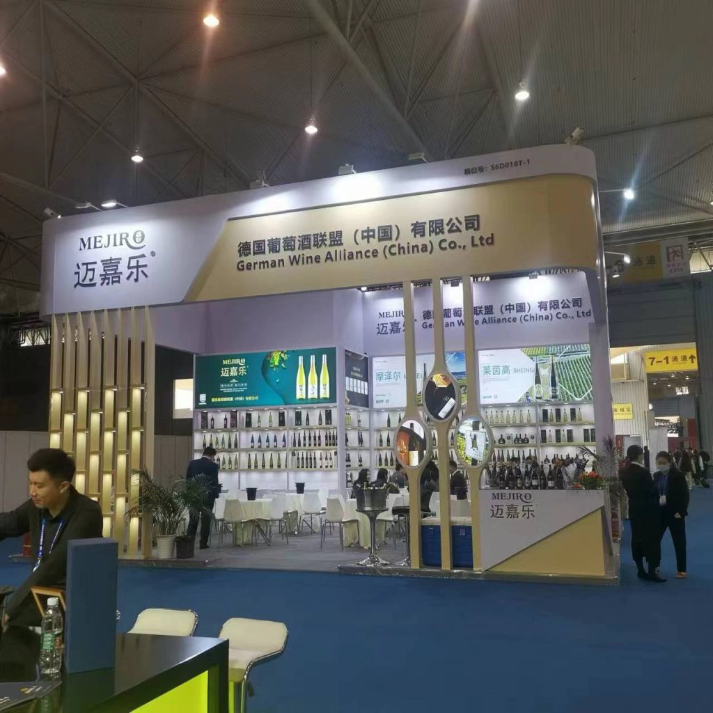 A busy trade show booth with numerous wine bottles displayed on shelves. Guests sit at small tables, and a large sign with text is visible overhead.