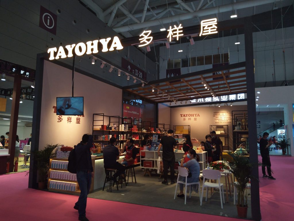 Crowded exhibition booth featuring products on shelves, a TV screen, and people interacting. The booth has a black frame and a pink carpet floor.