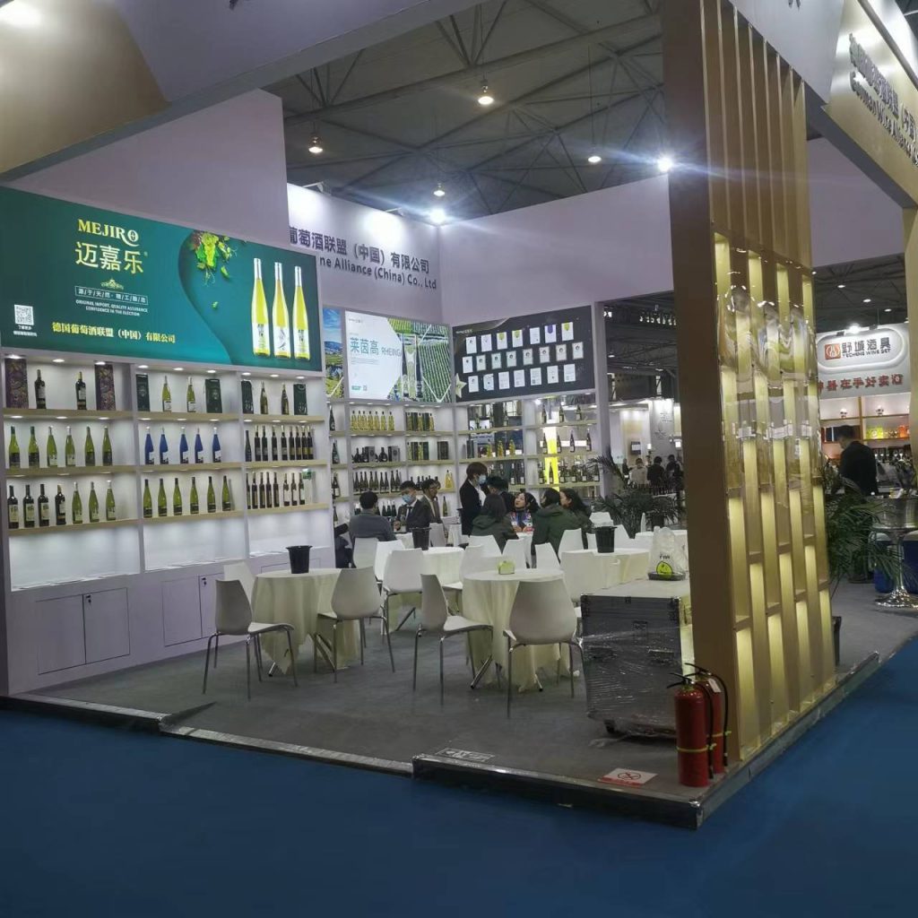 A wide shot of a trade show booth, featuring walls of wine bottles, small white tables with chairs, and a few attendees. A fire extinguisher is on the floor.