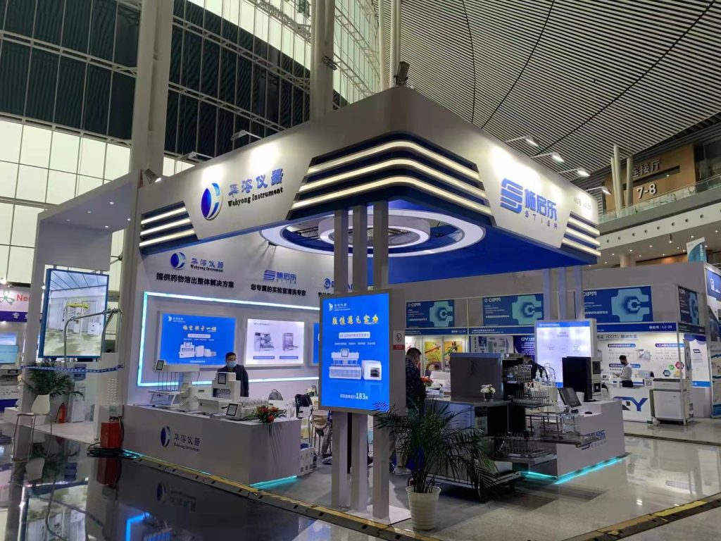 Exhibition booth with blue and white lighting, showcasing products and screens, with people interacting inside a large convention hall.