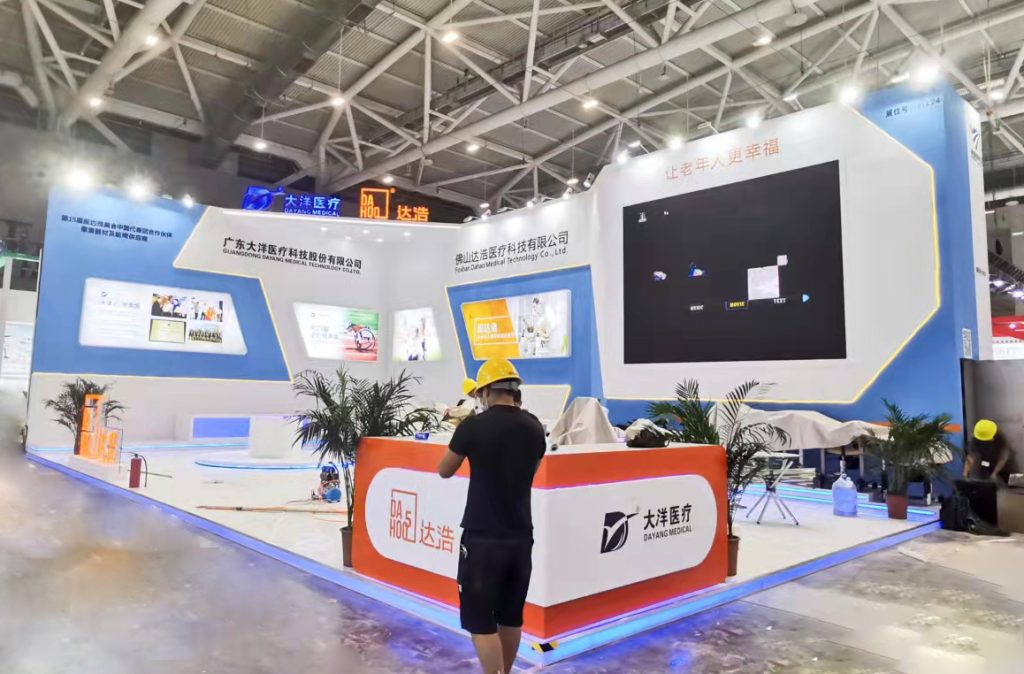Exhibition booth during setup, with palm trees, screens, and a worker in a hard hat.