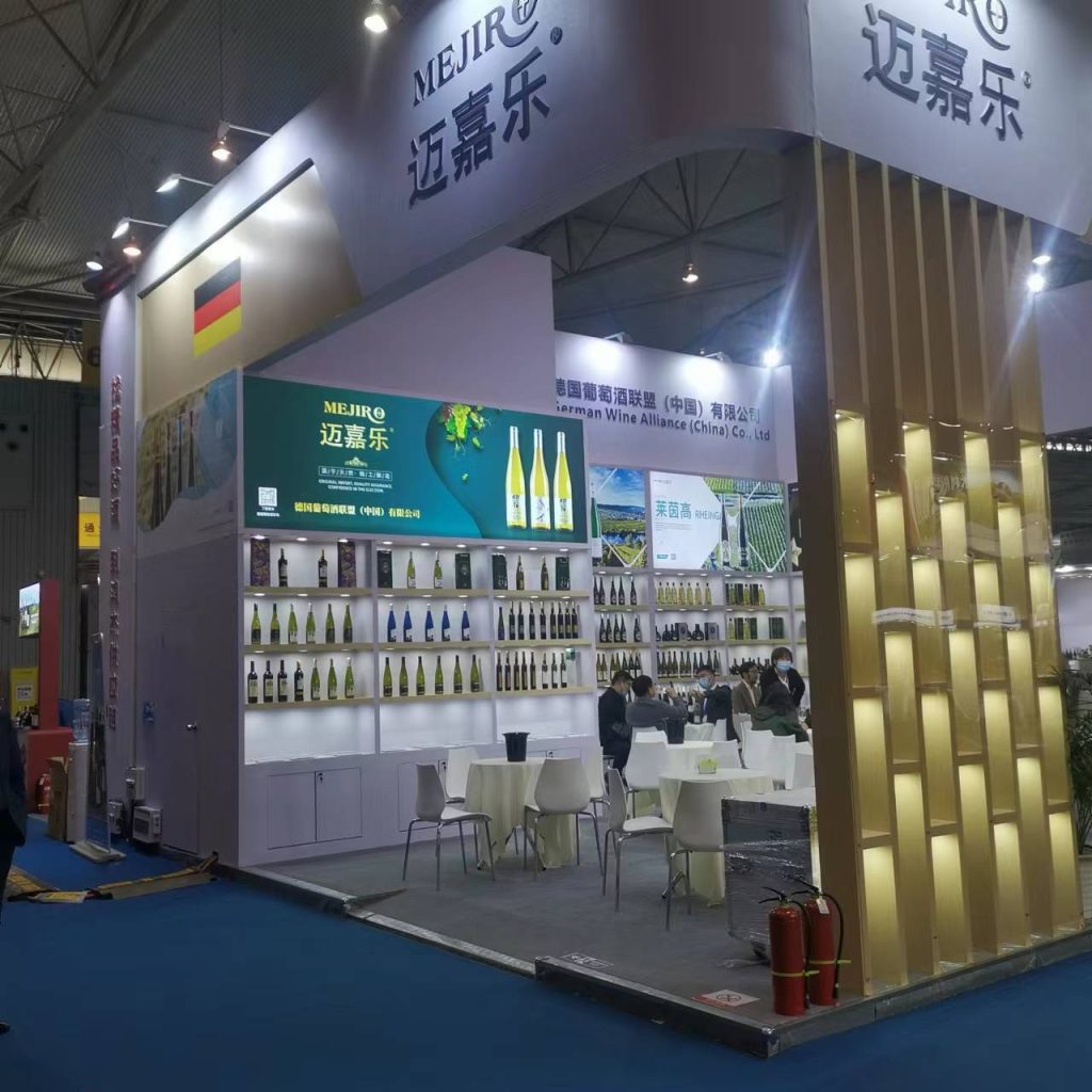 A trade show booth displaying rows of wine bottles on illuminated shelves. Several guests are seated at white tables, and banners with text are visible.