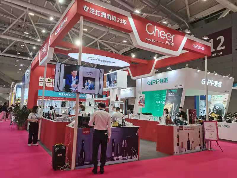 A busy red and white exhibition booth with products, a large video screen, and attendees walking past in a crowded convention hall.