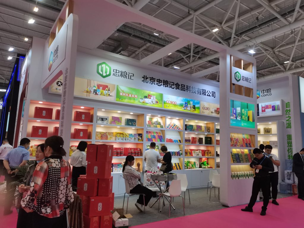 A busy exhibition booth with many people. White shelves display colorful packaged products, and a large screen shows food items. Some people are browsing, others are seated.