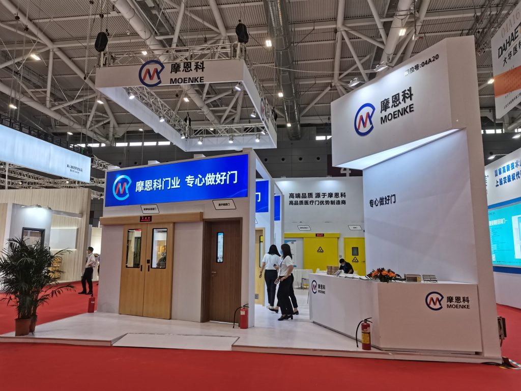An exhibition booth with multiple doors, a reception desk, and attendees browsing the display. A potted plant and red carpet are in the foreground.