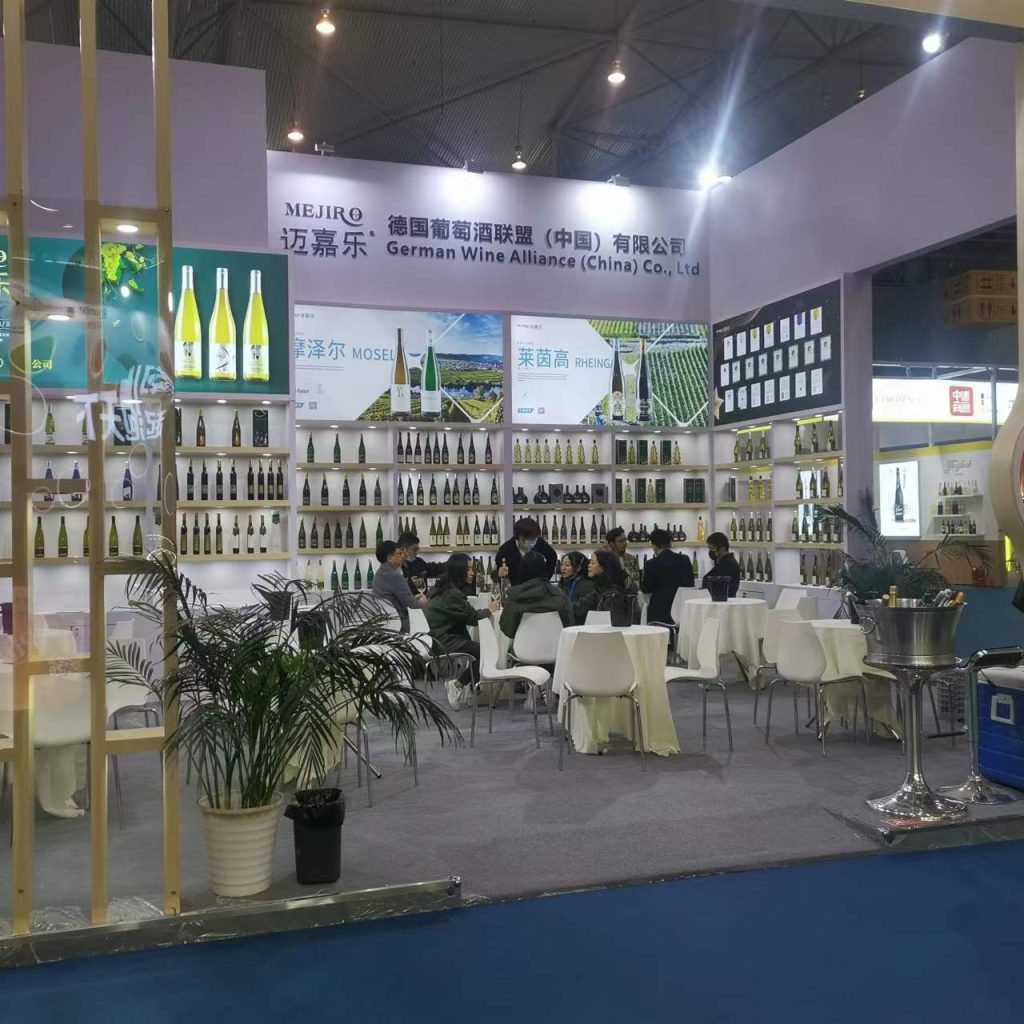 An interior view of a spacious trade show booth filled with shelves of wine bottles. Numerous white tables and chairs are set up for visitors, with plants decorating the space.