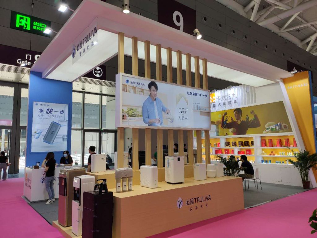 A modern exhibition booth displays various small appliances and water purifiers on wooden stands. A large screen features a man.