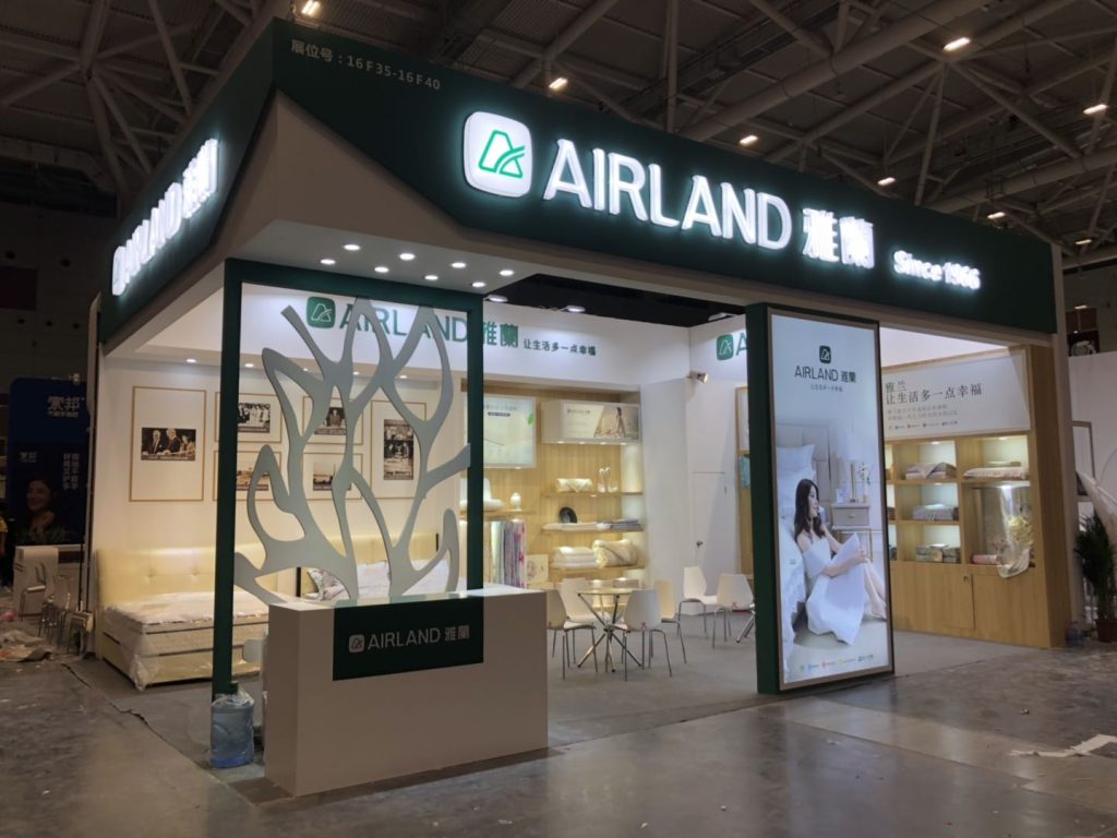 Trade show booth with bright signage, a bed display, and retail products. Construction materials and people are visible on the concrete floor.