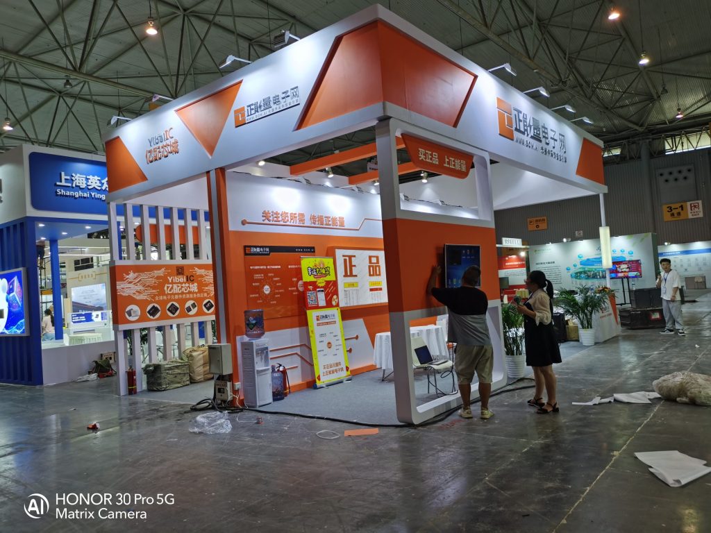 Two people setting up a white and orange exhibition booth in a large hall, adjusting a display screen.