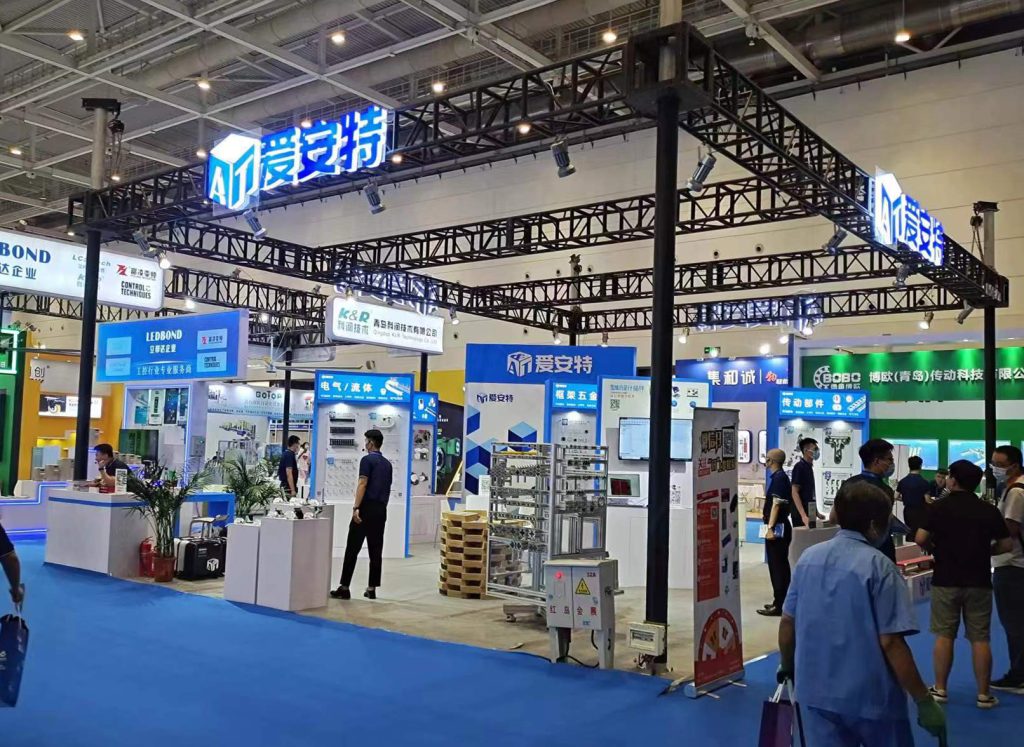 Bustling trade show floor with a booth featuring blue and white displays, an overhead metal truss with illuminated signs, and numerous visitors.