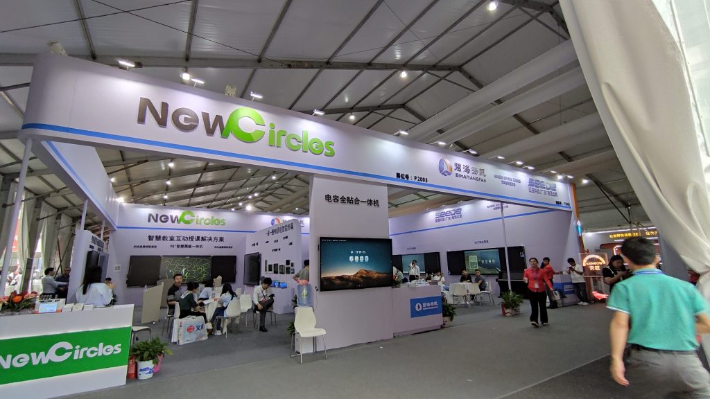 A busy technology trade show booth with multiple screens, informational displays, and attendees under a large tent structure.