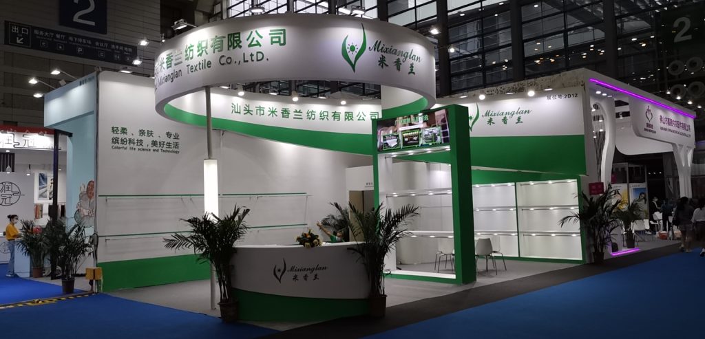 Real-world photo of a large green and white exhibition booth with plants, a reception desk, and large screens in an expo hall.