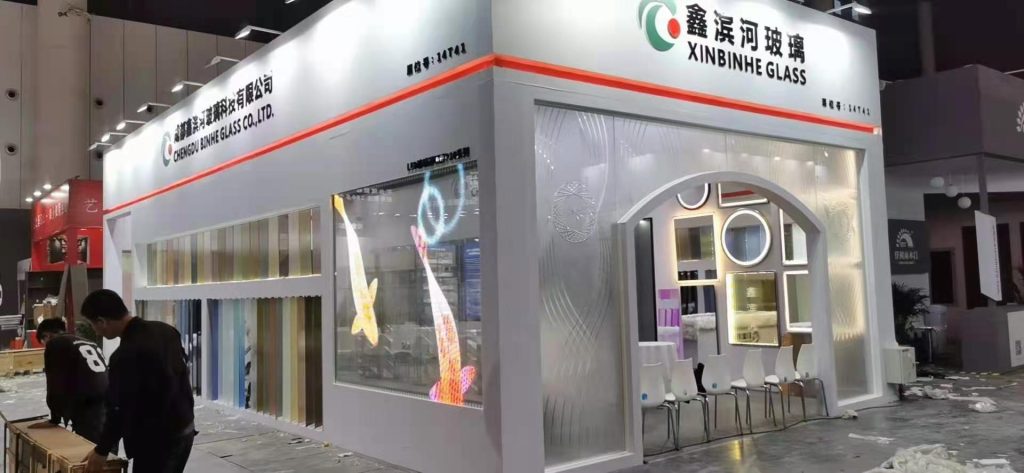 Exhibition booth displaying colored glass panels, a large transparent screen with two glowing fish, and an arched entrance with framed decor.