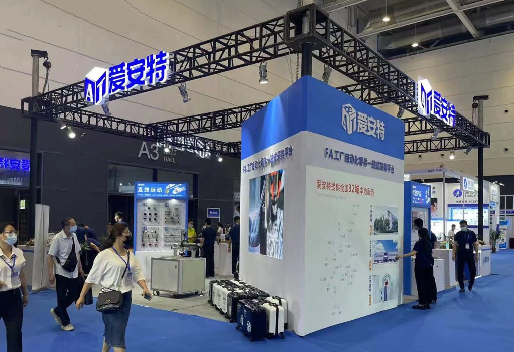 Trade show booth with a tall blue and white structure displaying graphics, under a metal truss. Attendees walk by the exhibit.