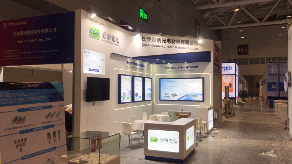 An exhibition booth within a large hall, featuring digital displays, a reception desk, seating, and a glass display case with small items. Other booths are visible.