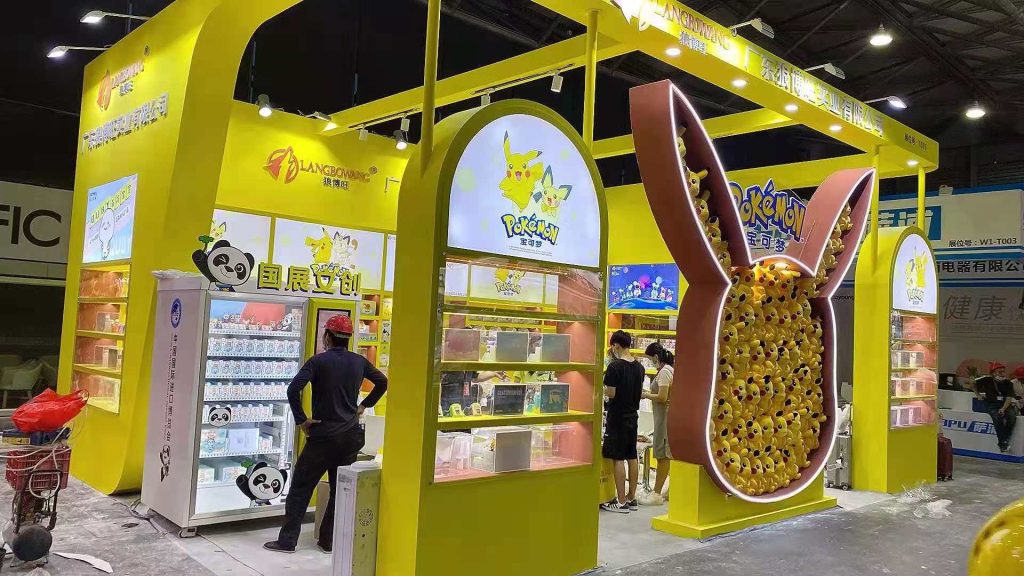 Yellow exhibition booth with a large, ear-shaped display full of yellow plush toys. Visitors stand near a refrigerator featuring an animated panda.