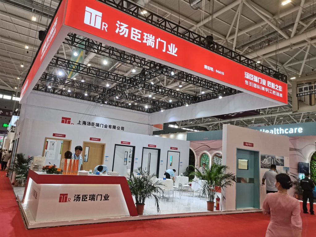 An exhibition booth with white walls and red overhead signs, displaying multiple doors. Several visitors and staff interact within the booth on a red carpet.