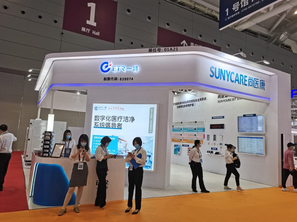 A brightly lit exhibition booth with an orange carpet, staff members interacting, and multiple display screens showcasing information.
