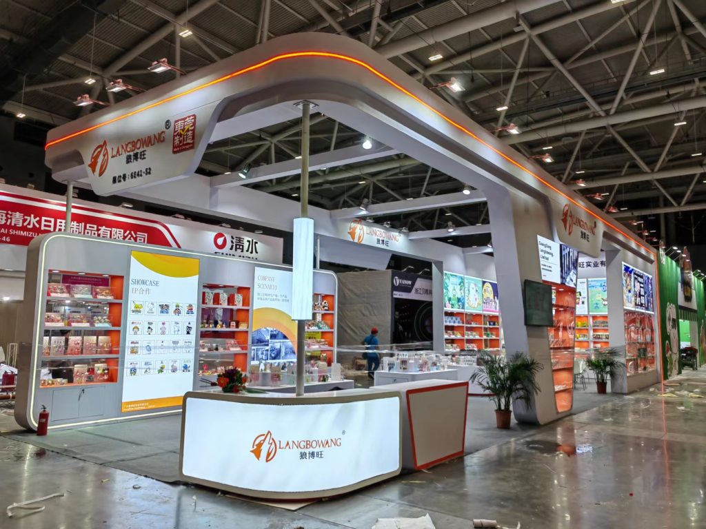 A modern exhibition booth in a large hall, featuring orange and white displays and a reception desk, with attendees visible.