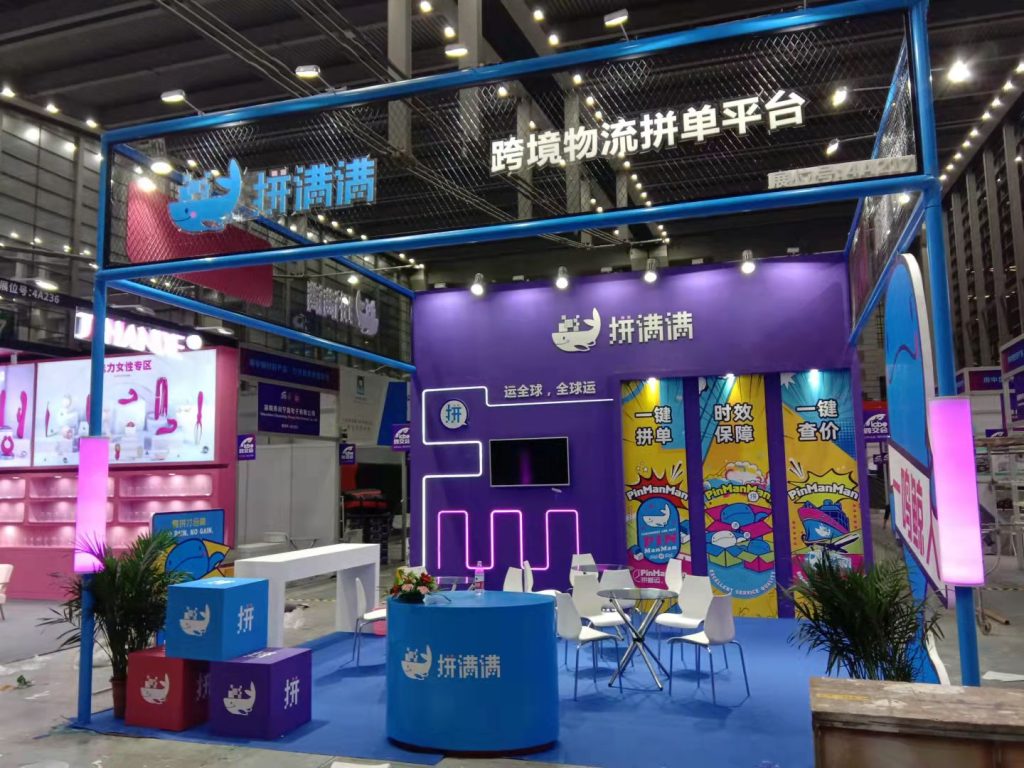 Real-world exhibition booth with purple walls and blue carpet, featuring a large screen, round tables, chairs, and character cutouts in a bright hall.