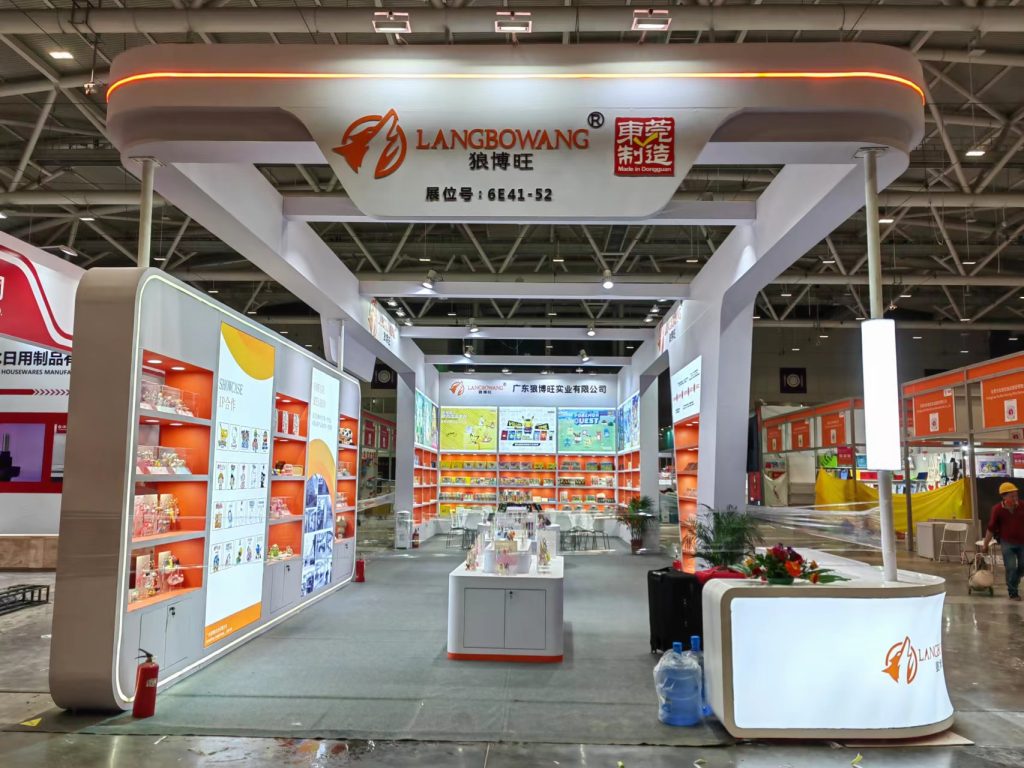 A wide view of a modern exhibition booth with orange and white displays, an overhead structure, and a reception counter in a convention hall.