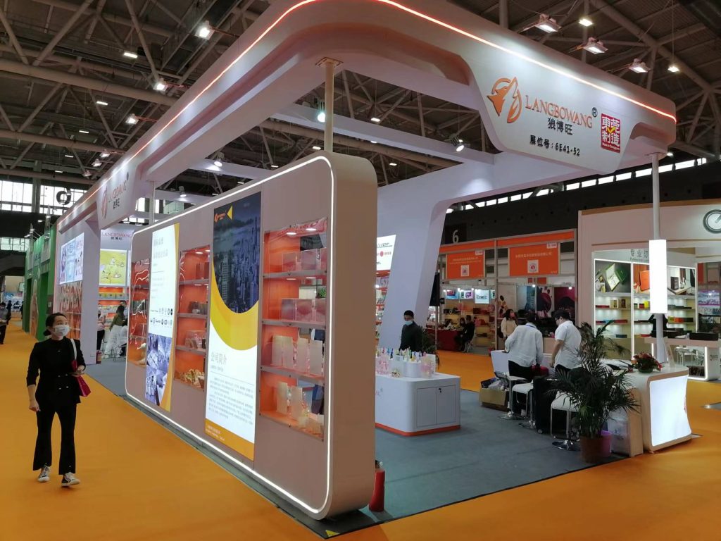 A person walks past a modern exhibition booth with illuminated orange shelves displaying products in a busy convention center.
