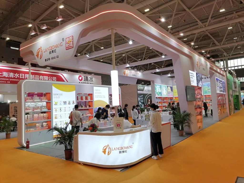 A bustling exhibition booth with attendees at display counters and a central desk, featuring modern orange and white aesthetics.