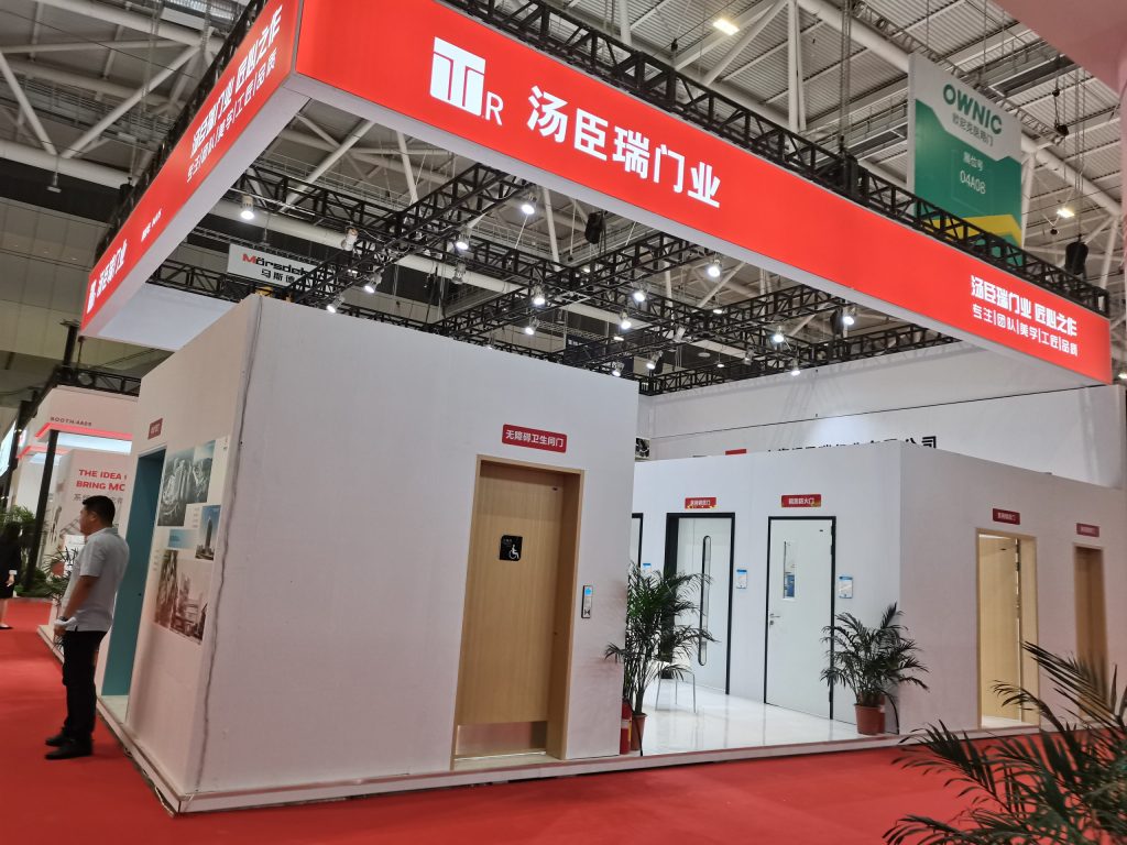 An exhibition booth with white walls and red overhead signs. It features various doors, including one with a wheelchair symbol. People are visible on the red carpet.