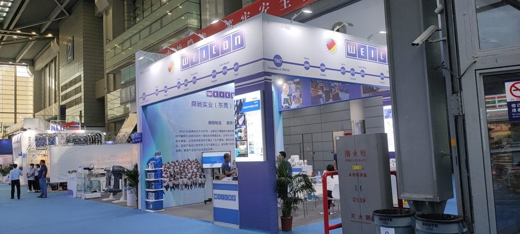 Attendees at a white and blue exhibition booth with large informational panels and plants.
