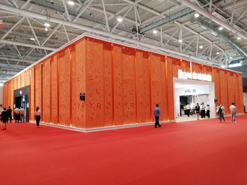 A large exhibition booth with orange walls featuring cutout letters and numbers, surrounded by a red carpet with people walking.