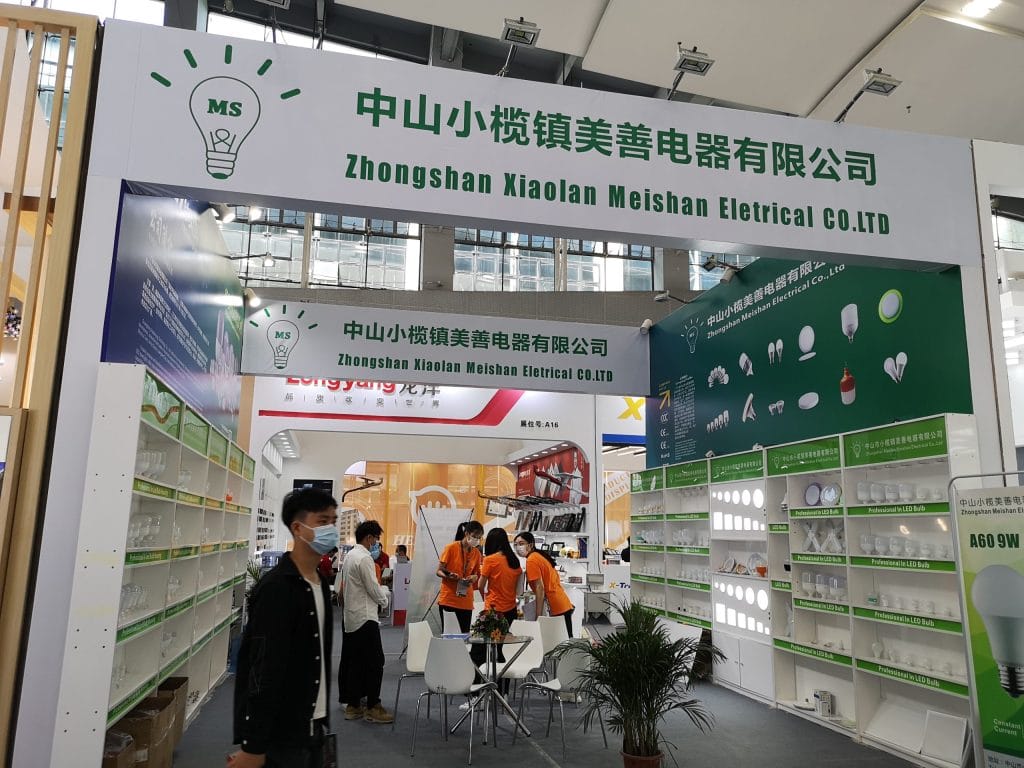 A busy exhibition booth with a green wall showcasing various light bulbs. Several people, some masked, interact with displays and products.