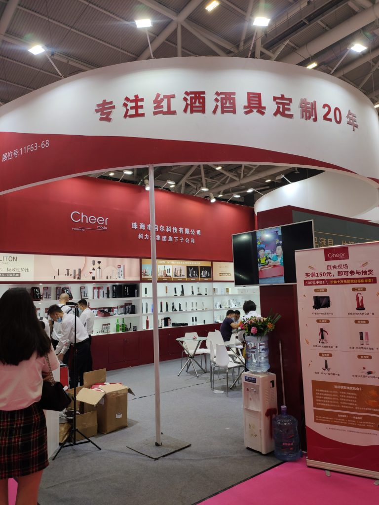 A closer view of an exhibition booth, showing people looking at displayed products. Red and white walls, shelves with bottles, a water cooler, and promotional banners are visible.