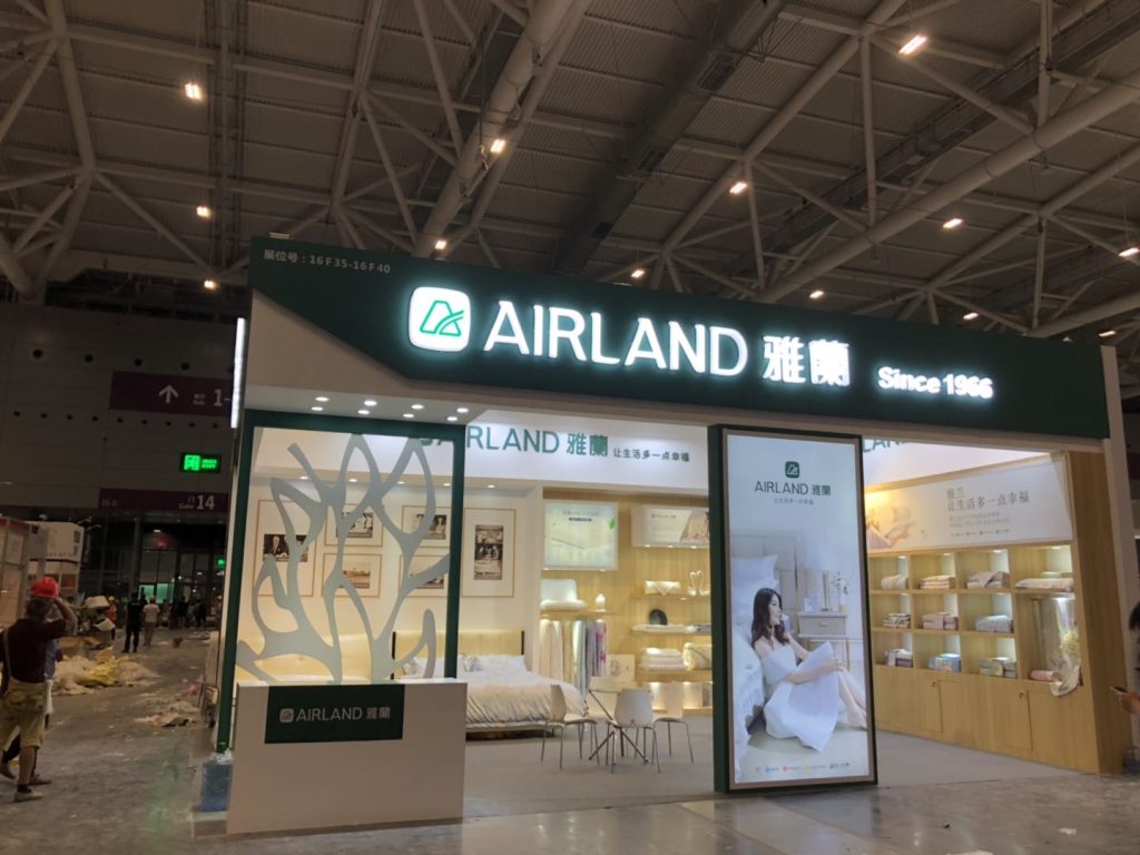 Brightly lit trade show booth featuring bedding, a bed display, and promotional posters. People and ongoing construction are in the background.