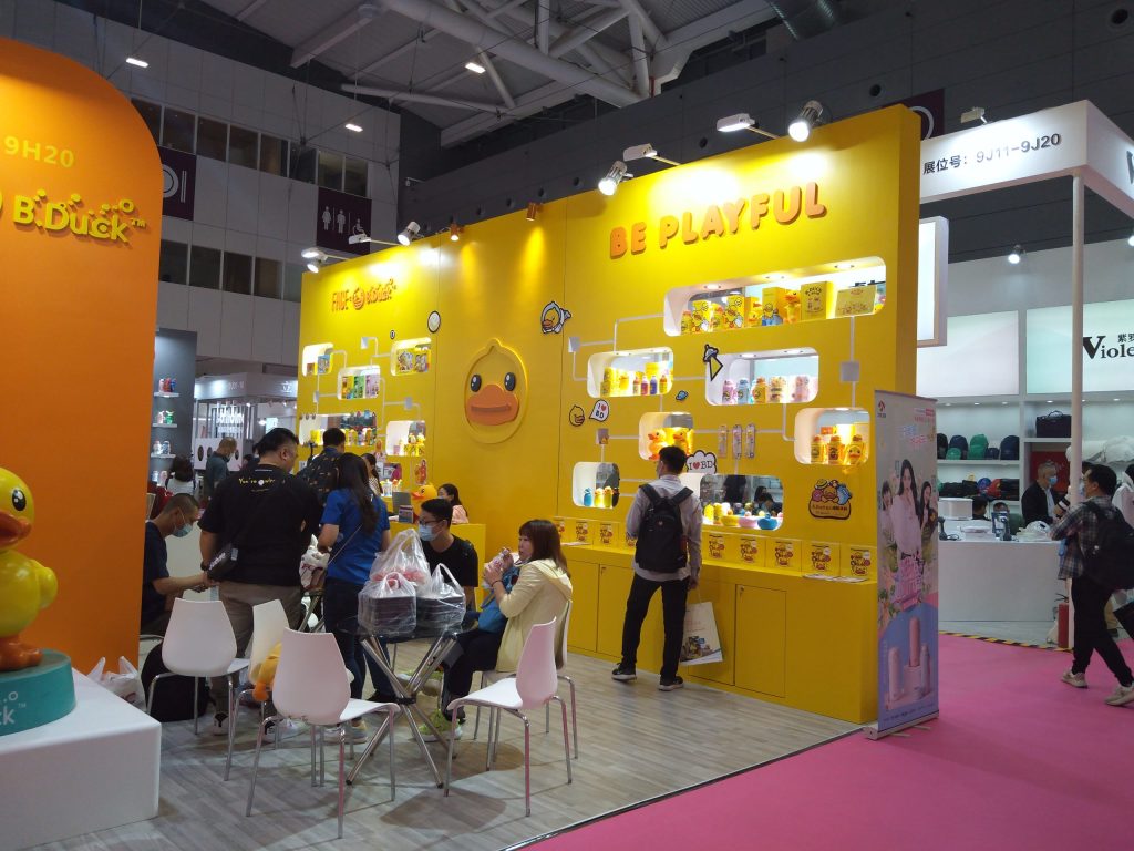 Visitors interact at a bright yellow trade show booth showcasing numerous products arranged on shelves, with some seated at tables.