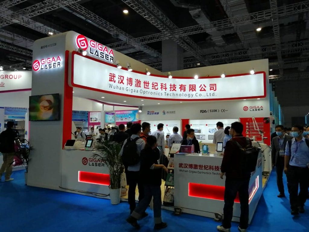A bustling exhibition booth with numerous people, illuminated displays, and a counter area on a blue carpet.